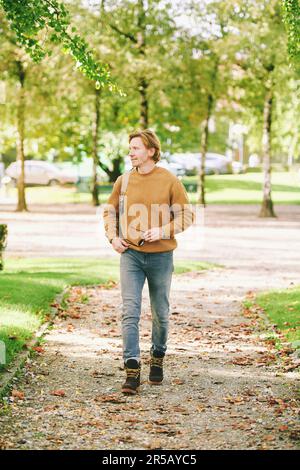 Portrait extérieur d'un jeune homme beau marchant à l'extérieur dans un parc public, portant un pull-over marron et un sac à dos Banque D'Images