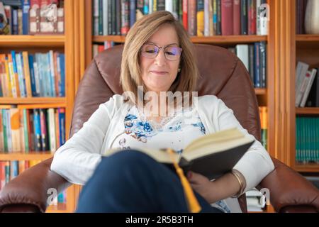 Femme blanche senior assise confortablement dans une chaise relaxante tout en lisant un livre. Banque D'Images