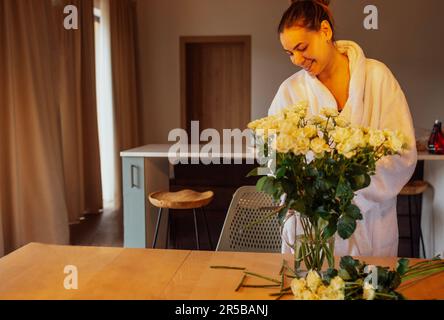 Jeune femme charmante en peignoir blanc coupe des tiges de roses blanches et les met dans un vase en verre. Souriant belle fille dans la chambre d'hôtel ou la cuisine dans elle Banque D'Images