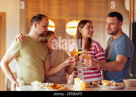 Deux couples mariés tiennent des verres de champagne et font des toasts dans la salle à manger à la maison. Les jeunes souriants dans des vêtements décontractés clink des gobelets avec des sparkli Banque D'Images