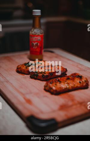 Trois côtelettes de porc fraîchement préparées sur une planche à découper en bois, accompagnées d'une bouteille de sauce chaude Banque D'Images