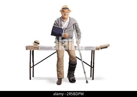 Patient âgé d'un homme dont le bras et la jambe sont cassés, assis sur un lit d'hôpital isolé sur fond blanc Banque D'Images