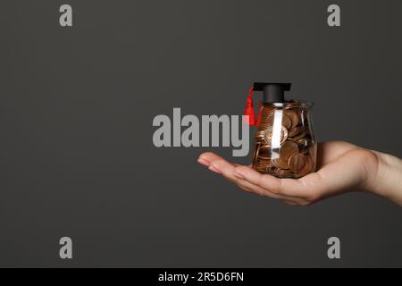 Femme tenant un pot en verre de pièces de monnaie et un chapeau de graduation sur fond gris foncé, gros plan avec un espace pour le texte. Concept de bourse Banque D'Images