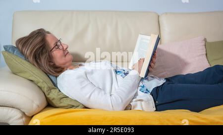 Femme blanche sénichée sur le canapé lisant un livre dans une attitude heureuse et détendue. Banque D'Images