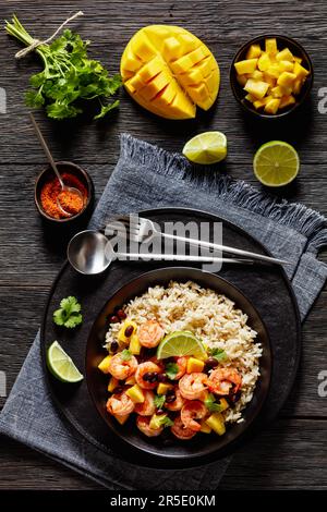 Crevettes grillées aux Caraïbes aux haricots noirs, salsa à la mangue et à l'ananas, servies avec du riz brun dans un bol noir sur une table en bois sombre, vue verticale Banque D'Images