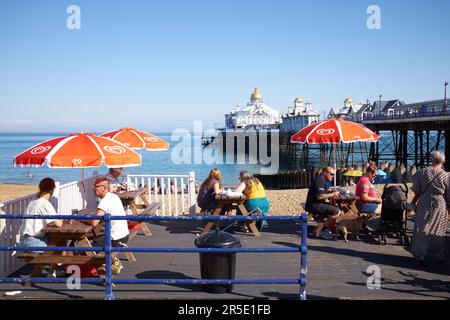 Eastbourne, East Sussex | Royaume-Uni - 4 août 2021 : les gens mangent et boivent sur des tables en terrasse avec le célèbre quai victorien en arrière-plan. Banque D'Images