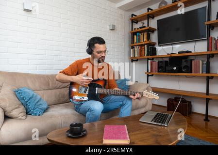 Jeune musicien débutant, débutant, portant un casque moderne, apprenant à jouer de la guitare et à enregistrer de la musique lors d'un cours de vidéo sur le Web à l'aide d'un ordinateur portable. Concentré millénaire Banque D'Images