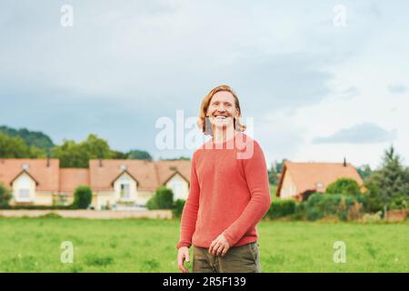 Portrait extérieur de modèle masculin heureux avec des cheveux rouges, posant dans une campagne avec de nombreuses maisons confortables sur fond Banque D'Images