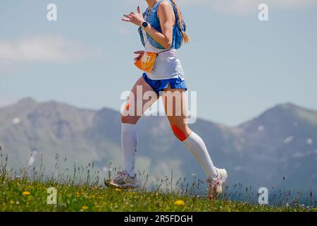 piste de course à pied d'athlète féminine sur un pré vert en arrière-plan de montagne, chaussettes de compression et genoux en kinésiotape Banque D'Images
