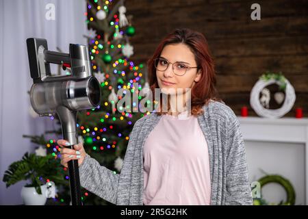 La jeune fille est debout, souriant doucement sur fond d'arbre de Noël et tenant un aspirateur dans la main. Banque D'Images