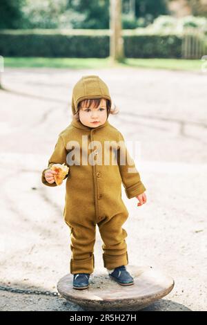 Portrait en plein air d'un petit enfant mignon s'amuser sur le terrain de jeu, porter un combinaison chaude, manger un croissant Banque D'Images