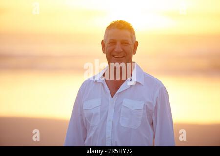 J'adore la plage. Portrait court d'un beau homme mûr debout sur la plage au coucher du soleil. Banque D'Images