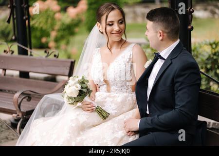 Le marié et la mariée sont assis sur le banc et se embrassent. Vue latérale de la femme élégante vêtue d'une robe de mariage Banque D'Images