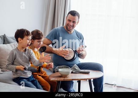 Papa joyeux s'amuser avec deux petits enfants dans le salon à la maison. Le jeune père joue le PAN comme une guitare. Les enfants adorables tiennent les cuillères à fente, rire Banque D'Images