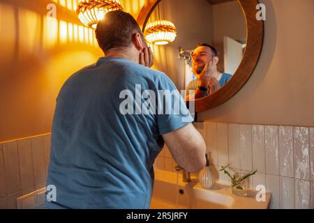 Jeune homme attrayant regarde la fenêtre et essuie ses mains avec une serviette blanche dans sa salle de bains. L'homme caucasien se lave le visage et les mains. hygie du matin Banque D'Images