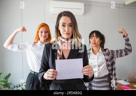 Trois femmes d'affaires au bureau, une femme au milieu tenant un papier avec écrit "Girl Power", ses collègues montrant biceps, concept d'autonomisation des femmes Banque D'Images