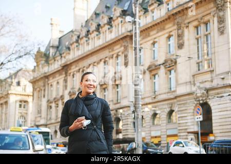 Bonne jeune femme marchant dans la rue, portant une veste chaude noire, tenant le téléphone et le masque facial dans les mains Banque D'Images