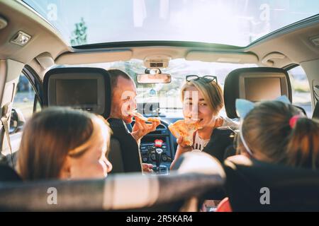 Jeune couple avec des filles mangeant une pizza italienne juste cuite dans une voiture moderne avec toit transparent. Des moments de famille heureux, l'enfance, la restauration rapide e Banque D'Images