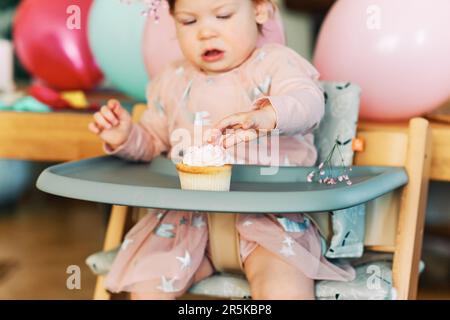 Adorable bébé fille de 1 ans mangeant un cupcake, enfant heureux assis dans une chaise, dégustation de dessert sucré, premier anniversaire Banque D'Images