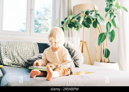 Adorable petit garçon assis sur un canapé et lisant ou regardant des photos dans un livre Banque D'Images