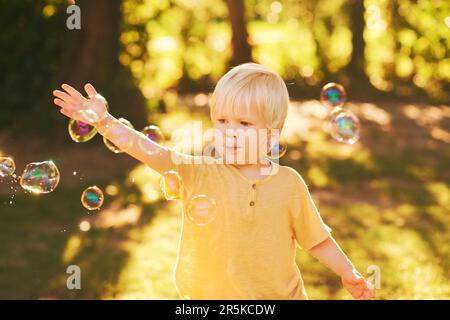 Portrait extérieur d'adorable petit garçon jouant avec des bulles de savon dans le parc d'été à la lumière du soleil, enfance heureuse et saine Banque D'Images