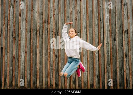 Portrait extérieur de la jeune fille heureuse portant un sweat-shirt gris, posant sur fond marron, sautant, mode pour les enfants Banque D'Images