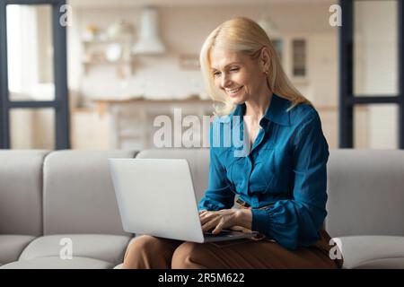 Une femme européenne mûre et heureuse utilisant un ordinateur portable et souriante, assise sur un canapé à la maison, travaillant en ligne, s'engageant dans la vie Internet Banque D'Images