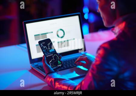 Vue de derrière une femme d'affaires moderne avec un ordinateur portable et un smartphone négociant des crypto-monnaies dans un bureau moderne. Banque D'Images