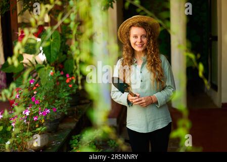 Jeune femme jardinier en chapeau de paille tenant la main pelle stands dans le jardin regardant la caméra sourire heureux. Femelle caucasienne gaie avec verger vert Banque D'Images