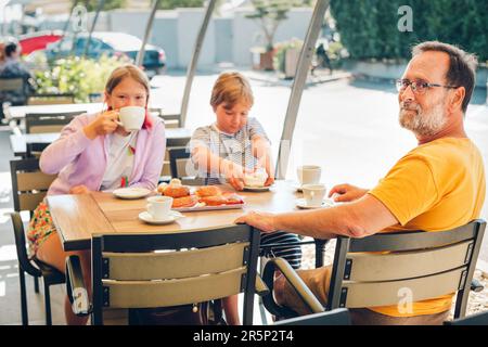 Famille avec enfants prenant le petit déjeuner dans un café ou un restaurant en plein air, boire du chocolat chaud, manger des boulangeries Banque D'Images