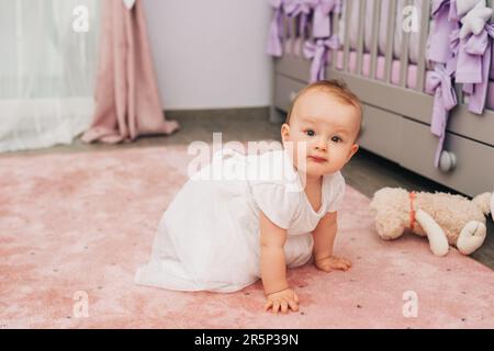 Portrait d'adorable fille de bébé qui se plie en pépinière, portant une robe blanche Banque D'Images