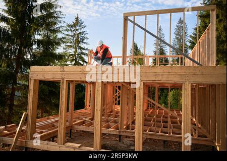 Carpenter construisant une maison en bois de deux étages près de la forêt. Homme barbu martelant des ongles dans la structure, portant un casque de protection, gilet de construction. Concept de construction écologique moderne Banque D'Images