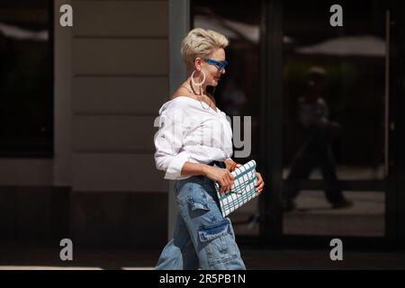 Femme sophistiquée avec des cheveux courts en chemise blanche et pantalon cargo jeans, accessoires tendance boucles d'oreilles, lunettes élégantes et marche d'embrayage extérieur Banque D'Images