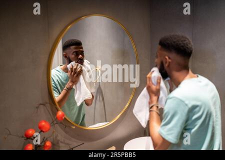 Un jeune homme ethnique africain mécontent essuie le visage après s'être lavé. Le mâle examine le miroir Banque D'Images