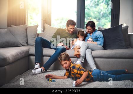 Nous avons du plaisir, peu importe ce que nous faisons. une mère, un père et leur fille utilisant une tablette ensemble sur le canapé tandis que leur fils joue sur le sol dans le Banque D'Images