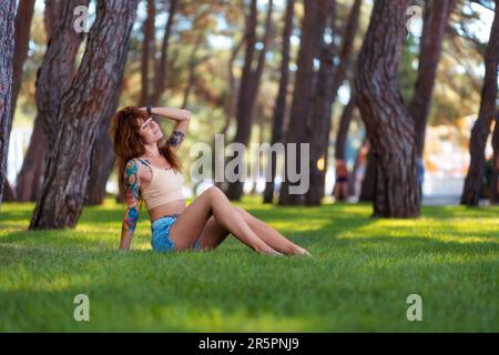 Caucasien belle jeune femme bronzée avec des tatouages posant sur la pelouse dans un parc bosquet. Vacances d'été en plein air. Concept de psychologie et de liberté. Banque D'Images