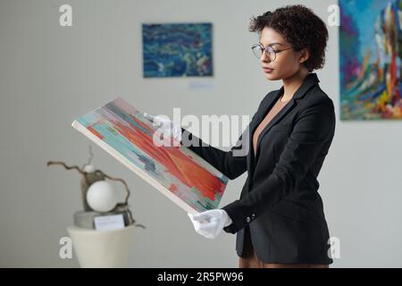 Jeune propriétaire de galerie en gants blancs tenant la peinture dans ses mains tout en travaillant dans la galerie Banque D'Images