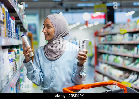 Une jeune belle musulmane dans un hijab choisit un produit pour se laver les cheveux, un shampooing, un après-shampooing, une femme tient un téléphone dans ses mains et lit les commentaires des clients en ligne dans un supermarché. Banque D'Images