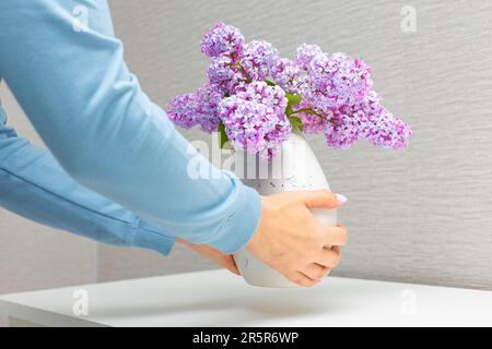 une femme met un vase avec une fleur de lilas sur la table. femme décorant la chambre avec des fleurs. mains tenant un vase de fleurs Banque D'Images