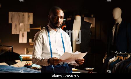 Homme tailleur vérifier les croquis dans l'atelier, en utilisant des dessins de vêtements et de tissu pour créer de nouveaux dessins de mode. Couturier professionnel travaillant dans un atelier sur mesure avec des outils industriels. Prise de vue à main levée. Banque D'Images