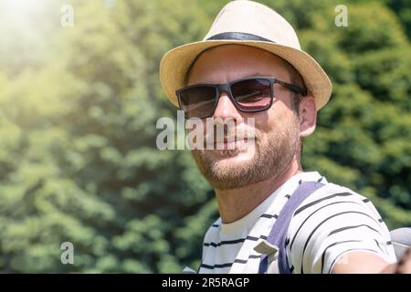 portrait selfie d'un homme adulte portant des lunettes de soleil et un chapeau de paille à l'extérieur en été. Banque D'Images