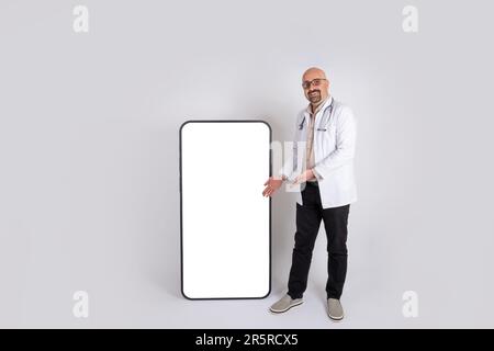 Médecin masculin présentant une nouvelle image d'idée de concept de publicité hospitalière. Photo complète du corps d'un médecin montrant un grand smartphone à l'écran vide, maquette. Banque D'Images