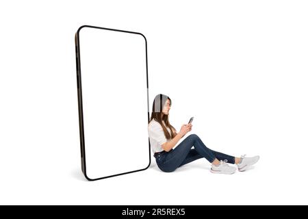 Fille caucasienne assise au sol avec téléphone sur sa main et smartphone bis penché. Écran blanc vierge énorme maquette de téléphone mobile géant. Banque D'Images