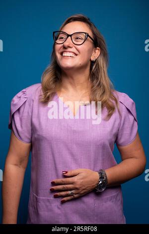 Jolie femme brésilienne portant des lunettes portant un uniforme d'infirmière lilas avec la main sur le ventre regardant vers le haut. personne positive. Isoald sur fond bleu. Banque D'Images