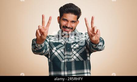 Jeune indien montrant le signe de la victoire, espérant le succès et la victoire, faisant le geste de la paix, souriant avec une expression aimable et optimiste. Beau type hindou barbu isolé seul sur fond beige studio Banque D'Images