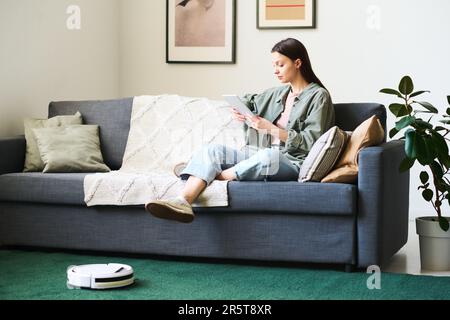 Jeune femme utilisant une tablette numérique tandis que le robot-aspirateur nettoie la moquette dans la chambre Banque D'Images