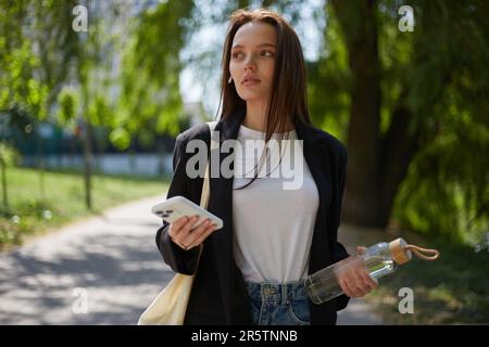 Belle jeune femme avec de longs cheveux bruns marchant dans le parc vert avec une bouteille d'eau en verre et un smartphone moderne dans les mains. Portrait d'un brett Banque D'Images