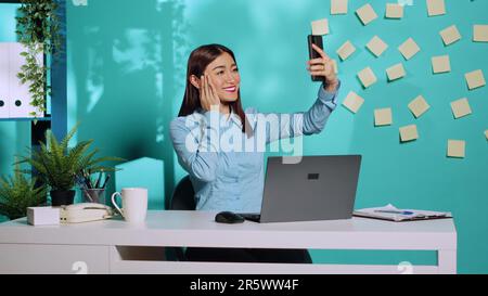 Joyeux employé pétillant prenant des selfies photo avec un smartphone, en lançant le signe de paix geste de main et en soufflant baiser à l'appareil photo. Femme d'affaires s'appréciant dans un bureau coloré et détendu Banque D'Images