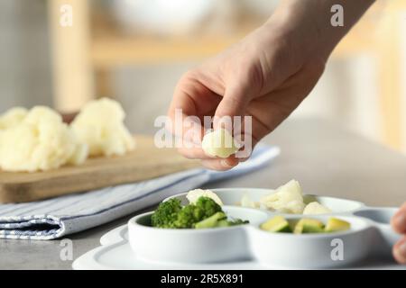 Nourriture pour bébé. Femme mettant le morceau de chou-fleur dans la section plaque avec différents légumes à la table grise texturée, gros plan Banque D'Images