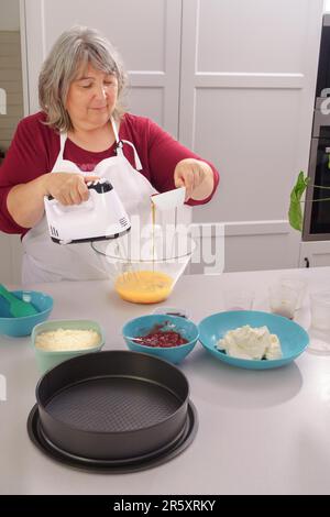 Femme chef dans un tablier blanc battant des oeufs dans un bol avec un mélangeur électrique pour faire un gâteau de fraise Banque D'Images